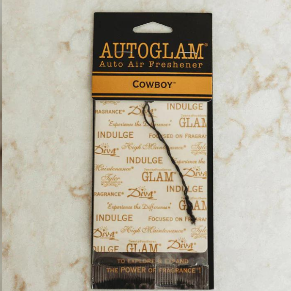 Commercial product photography of a Tyler Candle Company AUTOGLAM Cowboy air freshener resting on a marbled surface featuring a black and orange header card and a white tag adorned with a black hanging string and repeating gold Diva and High Maintenance text