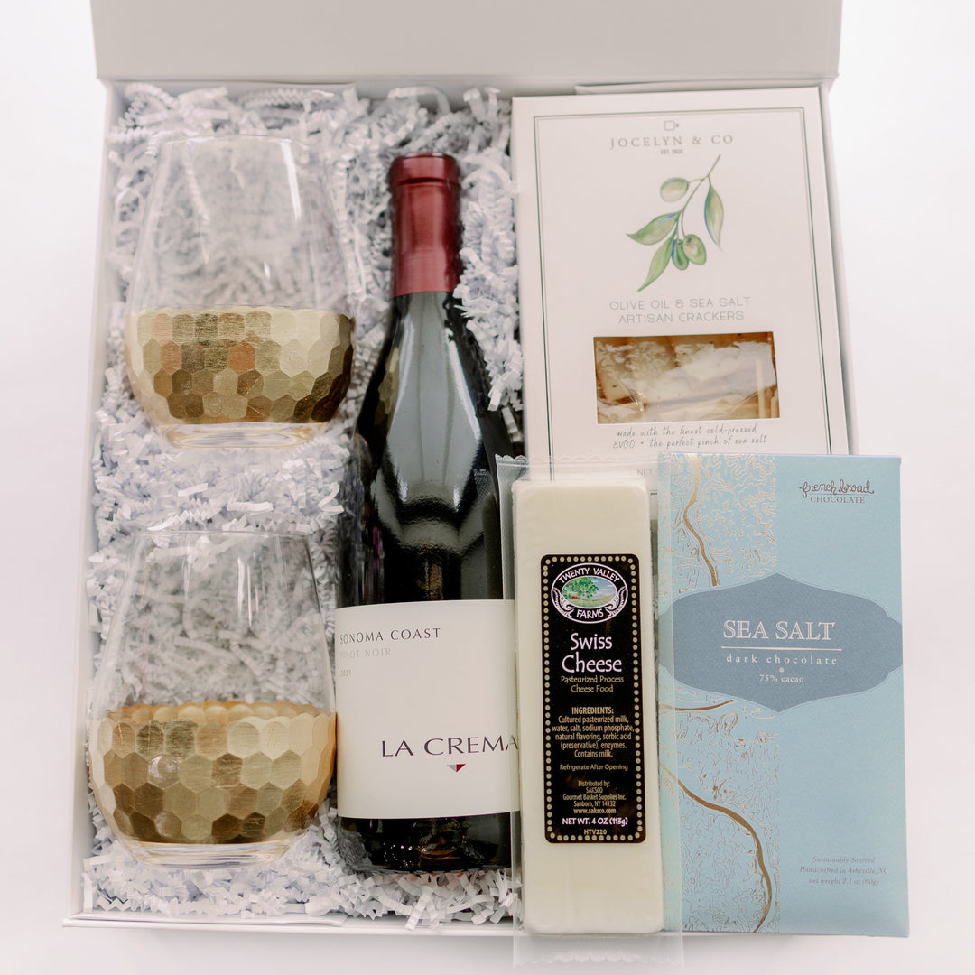 Digital photograph of a luxury wine gift set in a white presentation box containing a bottle of La Crema Pinot Noir, two gold honeycomb-patterned stemless glasses, and a selection of artisan cheese and crackers on white shredded paper