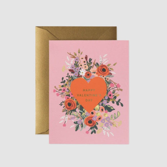 Illustrated greeting card by Rifle Paper Co featuring a large coral heart with gold foil lettering reading Happy Valentine's Day surrounded by a vibrant floral wreath of red roses and purple flowers on a light pink background paired with a kraft paper envelope
