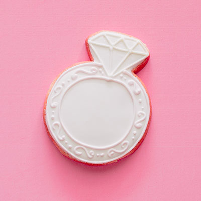 White-iced sugar cookie shaped like a diamond engagement ring on a pink background