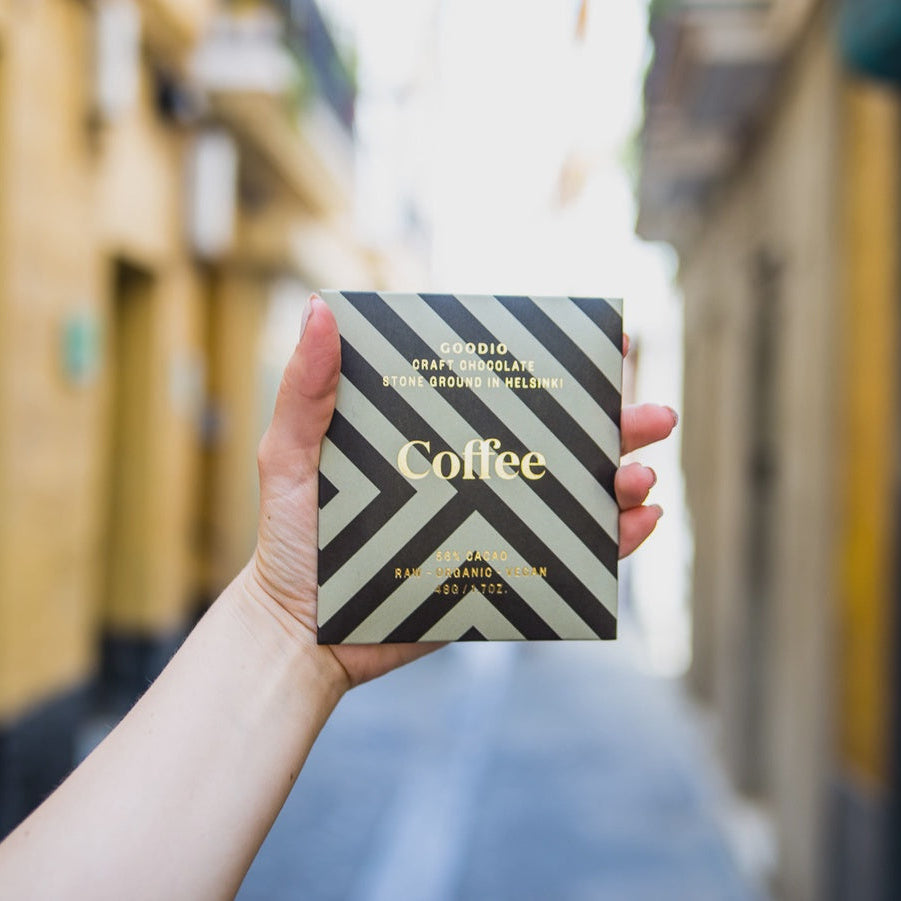 Digital photograph of a hand holding a Goodio Coffee craft chocolate bar featuring a sage green geometric chevron pattern and gold lettering against a blurred sunlit narrow European cobblestone street