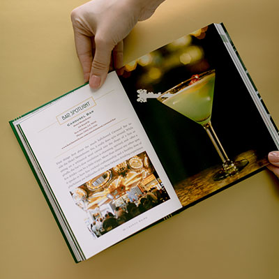 Photograph of an open book detailing the iconic Carousel Bar at Hotel Monteleone in New Orleans featuring a pale green martini with a white key-shaped garnish and a vibrant photo of the ornate gold-lit revolving bar