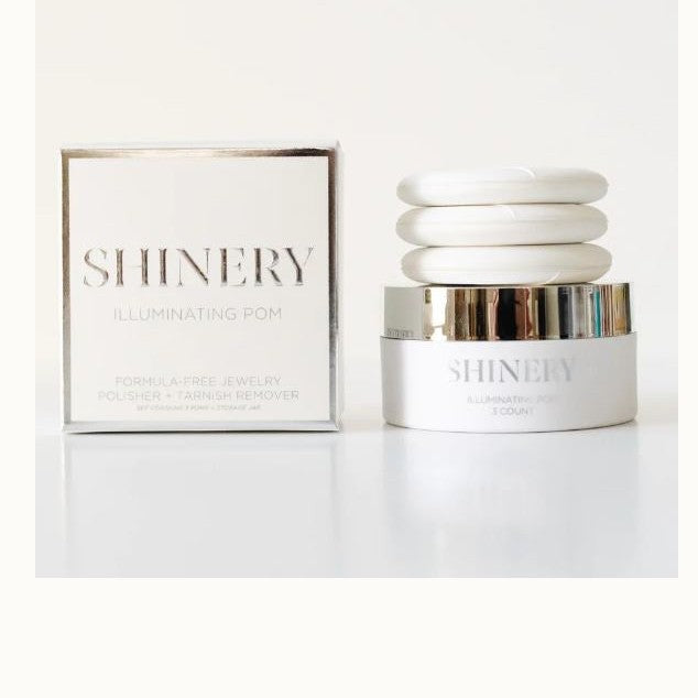 Studio product photography by Shinery showcasing the Illuminating Pom jewelry cleaning kit featuring a white square box with metallic silver-foil text next to a jar topped with three stacked circular white pads on a minimalist white surface