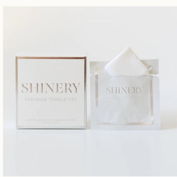 Product photograph by Shinery of luxury jewelry cleaning wipes featuring a square white box with rose gold foil lettering and an open individual sachet revealing a folded white towelette on a reflective white background