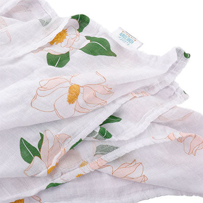 Cotton muslin swaddle blanket by Little Hometown featuring a southern magnolia floral pattern with delicate pink blossoms, vibrant green leaves, and textured golden-yellow centers