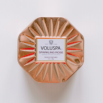 Commercial product photograph of a Voluspa Sparkling Rose candle featuring an octagonal rose gold tin with an embossed sunburst texture and a white square label on a neutral background